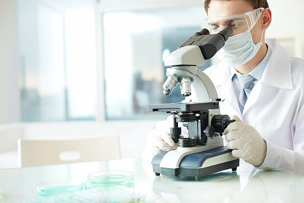 Laboratory employee wearing a face mask, gloves and safety goggles in a white lab coat sits in the lab and looks through a microscope.